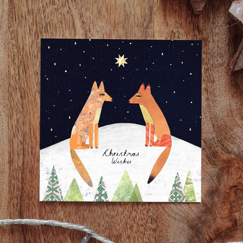Collage style illustration with two foxes facing each other on top of a snow covered hill. There are Christmas trees in front of the front, and a star filled night sky in the background. There is a bright yellow star in the middle of the sky between the foxes. 