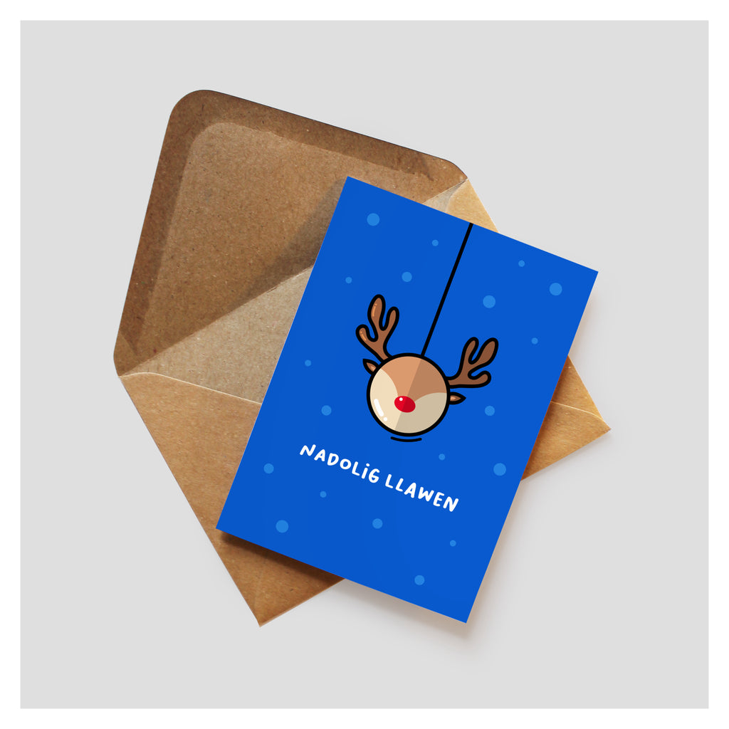 Blue background with snow falling. A graphic minimal reindeer head shaped bauble hangs in the centre, with 'Nadolig Llawen' underneath in white text. The card rests on top of a brown kraft envelope. 