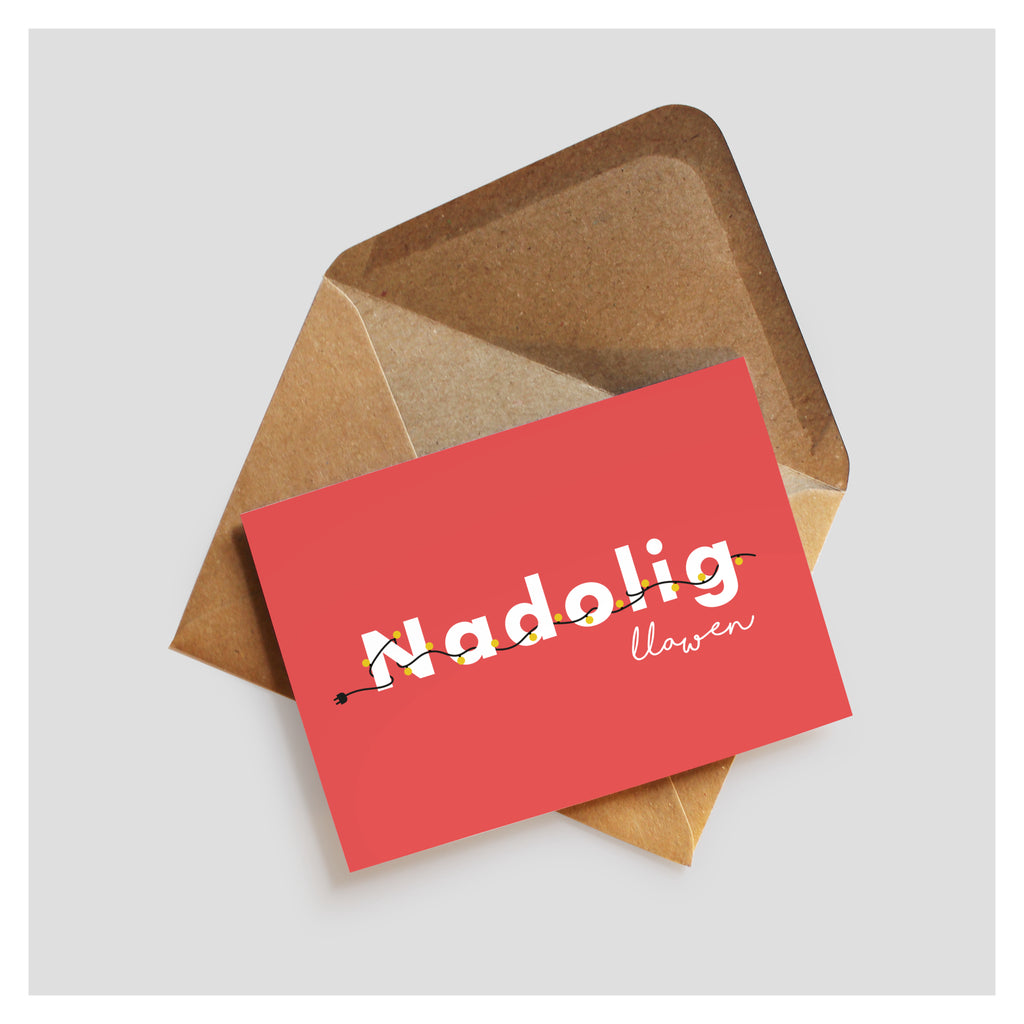 Red greeting card with 'Nadolig Llawen' is written across in white font. Woven in and out of the 'Nadolig' is a string of christmas lights with yellow bulbs. The card rests on top of a brown kraft envelope.