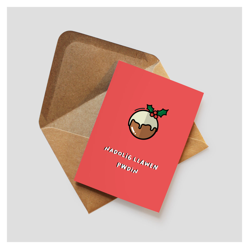 Red greeting card with cartoon style illustration of a Christmas pudding with the text 'Nadolig Llawen Pwdin' underneath. the card rests on top of a brown kraft envelope.