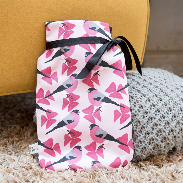 Lifestyle image.  Pink patterned fabric with bullfinch motif hot water bottle with black ribbon.