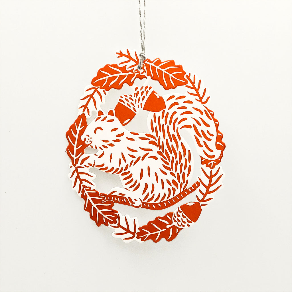 The image shows a squirrel sat on a branch below acorns, the squirrel is framed by an oak leaf garland. The image is embossed onto white paper and the image highlighed with copper coloured foil. The decoration is cut-out and hung from a thread. 