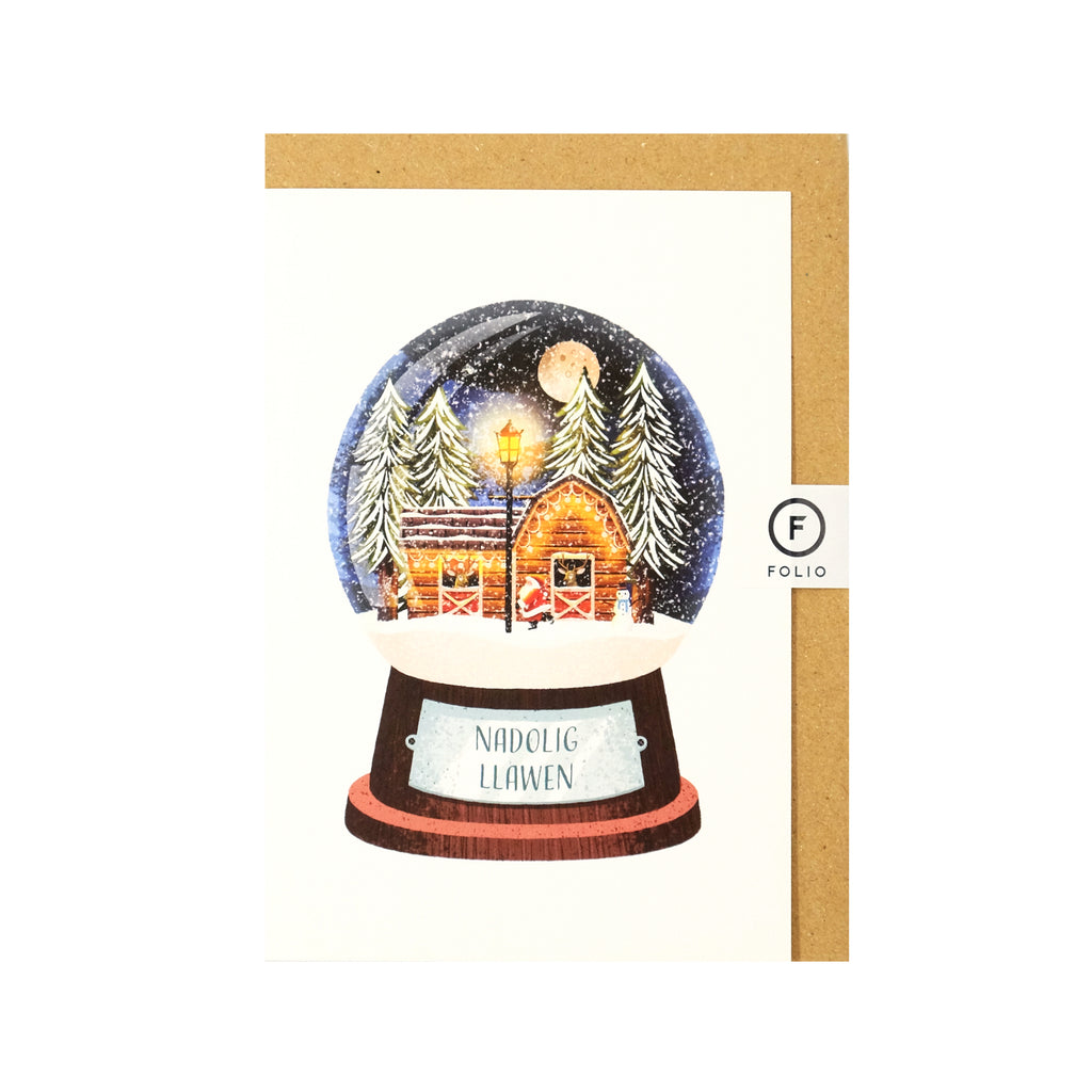 Welsh language Christmas card featuring an illustration of a snowglobe. Inside the globe is a wintery night scene of Santa tending to his reindeer who are in a log cabin with large snowcapped christmas trees behind the cabin There is a big full moon in the sky and a lampost and snowman in front of the cabin. The plaque on the globe reads "nadolig Llawen"