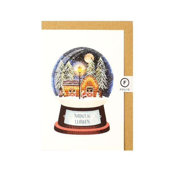 Welsh language Christmas card featuring an illustration of a snowglobe. Inside the globe is a wintery night scene of Santa tending to his reindeer who are in a log cabin with large snowcapped christmas trees behind the cabin There is a big full moon in the sky and a lampost and snowman in front of the cabin. The plaque on the globe reads "nadolig Llawen"