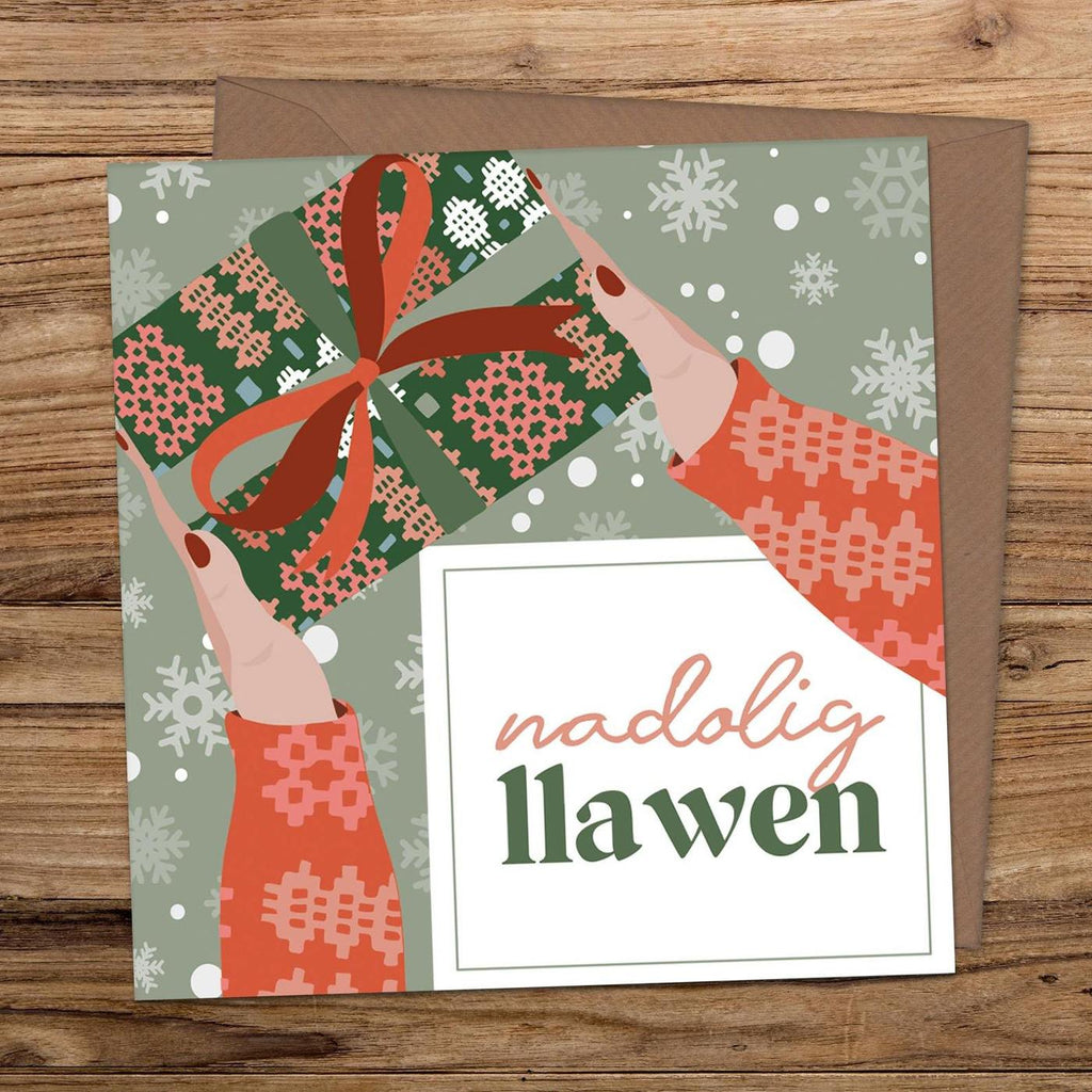 Illustrated Christmas card showing two hands with the sleeves of a festive welsh blanket pattern jumper showing. They are handing over a present wrapped in green and pink welsh blanket style wrapping paper and finished with a red bow.
