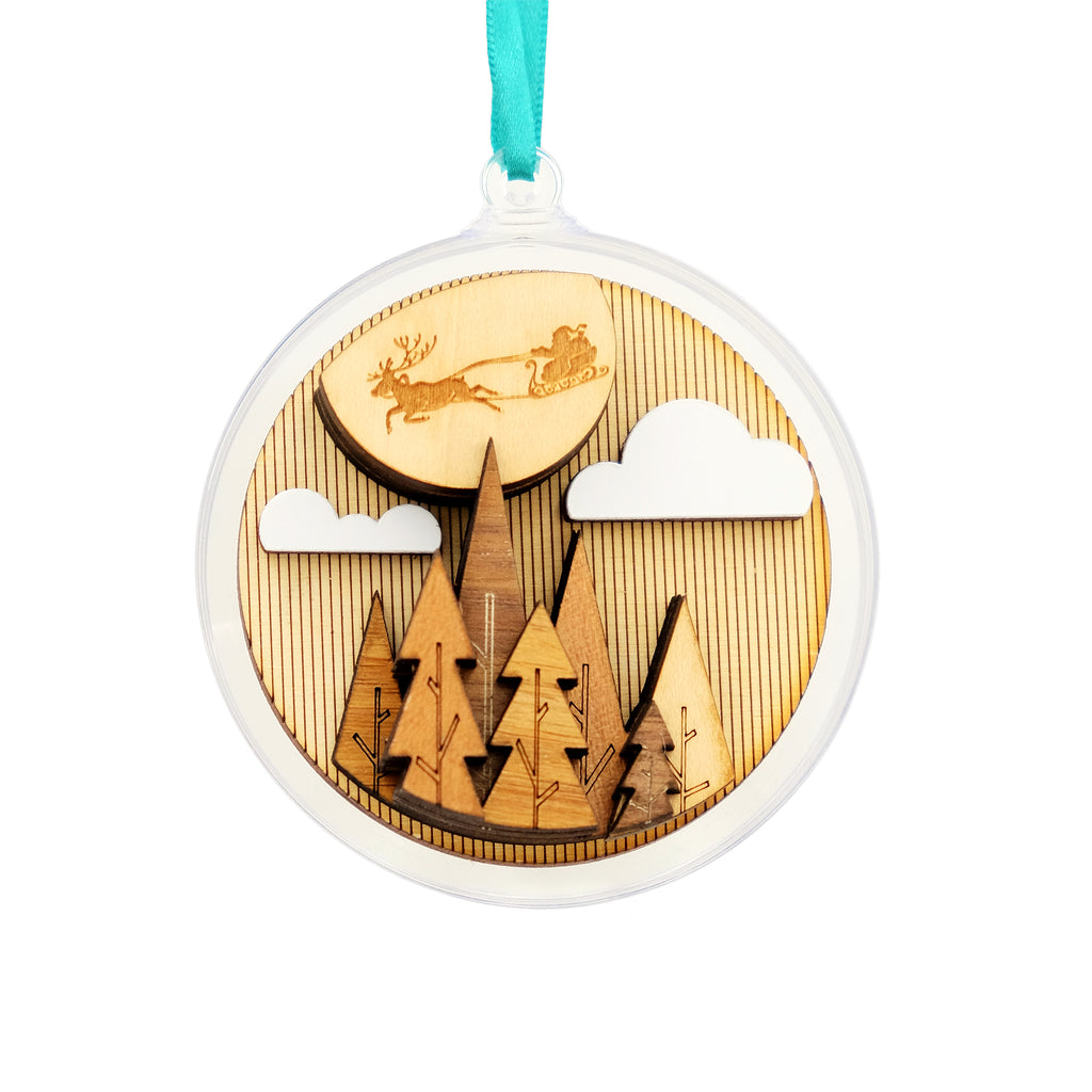 The image shows a festive scene of santa in his sleigh flying across the moon amongst some clouds. There are trees below. The scene is crafted from laser cut wood in various shades, with the clouds painted white. The wood is suspended in an acrylic bauble and hung from a pale coloured ribbon. 