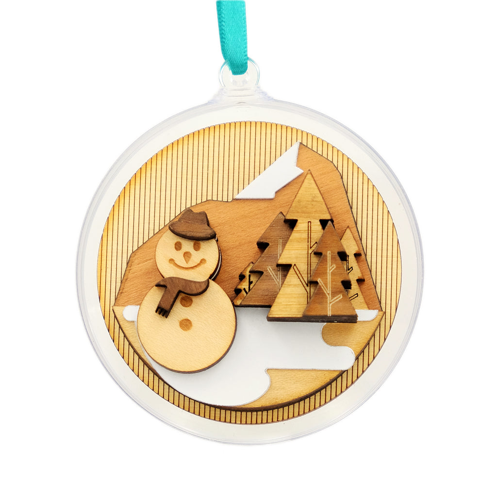 festive scene made from layers of laser cut wood. The scene shows a snowman in front of trees and mountains. The wood is suspended in an acrylic bauble and hung from a satin ribbon 