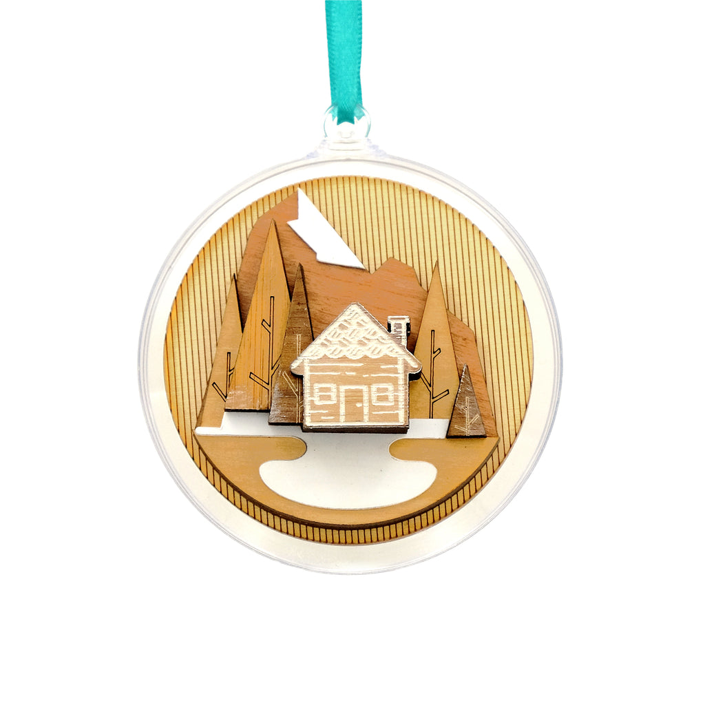 This festive scene is made from layers of laser cut wood in varying tones. It shows a gingerbread style cabin in front of trees and snowcapped mountains. The wood is suspended in an acrylic bauble and hung from a satin ribbon.