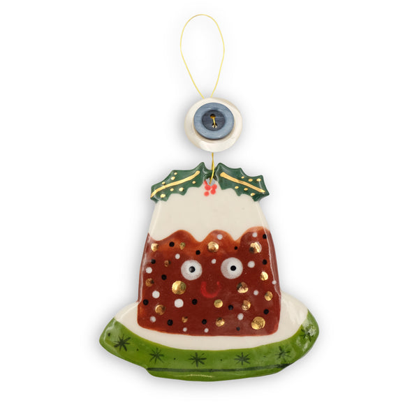 Ceramic Christmas Hanging Decoration. The pudding sits on a green and white plate, it has icing and a sprig of holly and berries on top. The pudding as eyes and a smile. It hangs from gold coloured wire with vintage buttons on them. 
