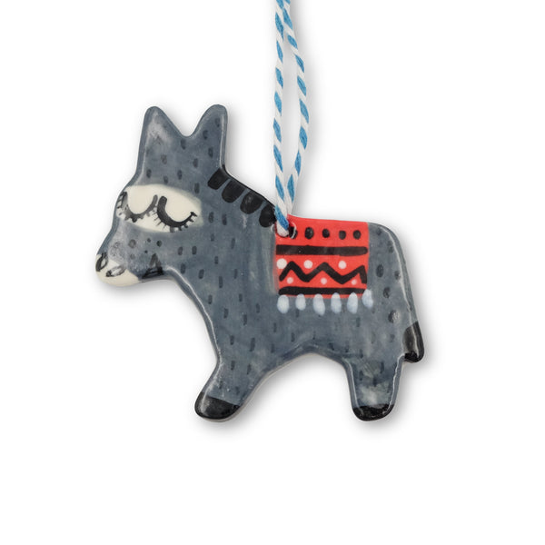 Ceramic hanging decoration. Grey donkey in side profile with a red blanket draped over its back. It hangs from blue and white twine. 