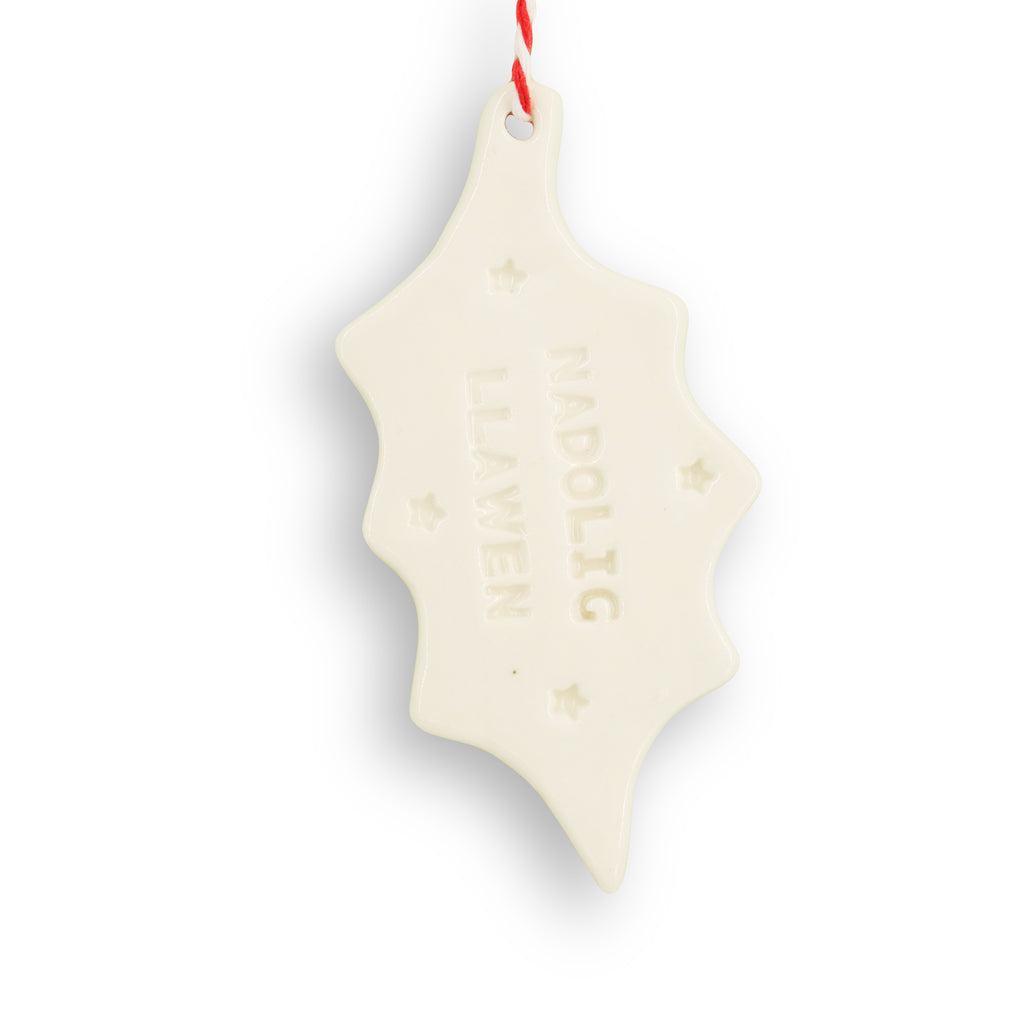 White ceramic hanging decoration. Holly leaf shape, with "Nadolig Llawen' stamped into the surface along with small stars. It hangs from red and white twine. 