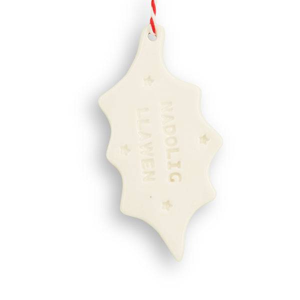 White ceramic hanging decoration. Holly leaf shape, with "Nadolig Llawen' stamped into the surface along with small stars. It hangs from red and white twine. 