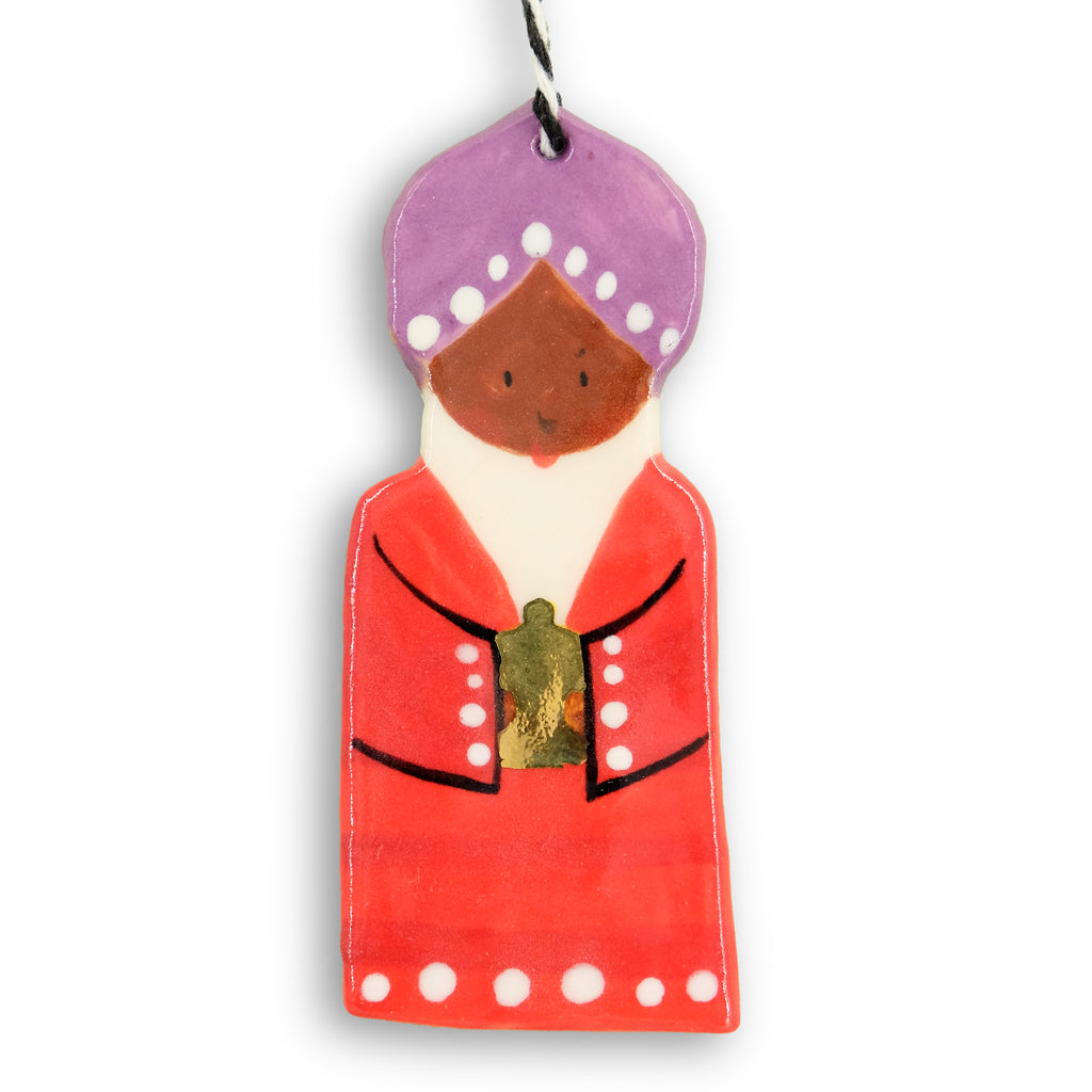 Ceramic hanging decoration. Wiseman holding a gold bottle. Wearing a purple turban and dark orange robe with white dots. 