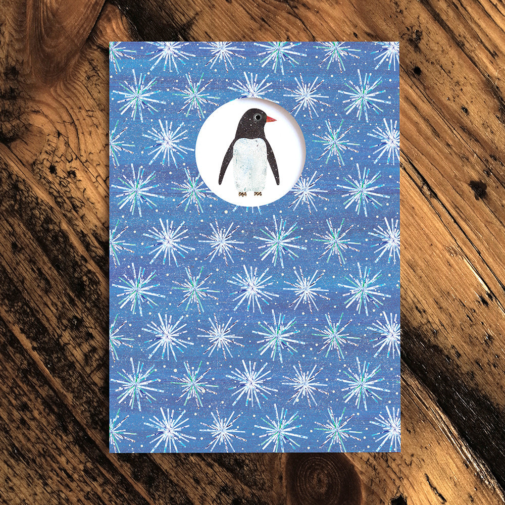 Christmas card with a pale purple background and white snowflake pattern. White circle cut out with a penguin illustration. 