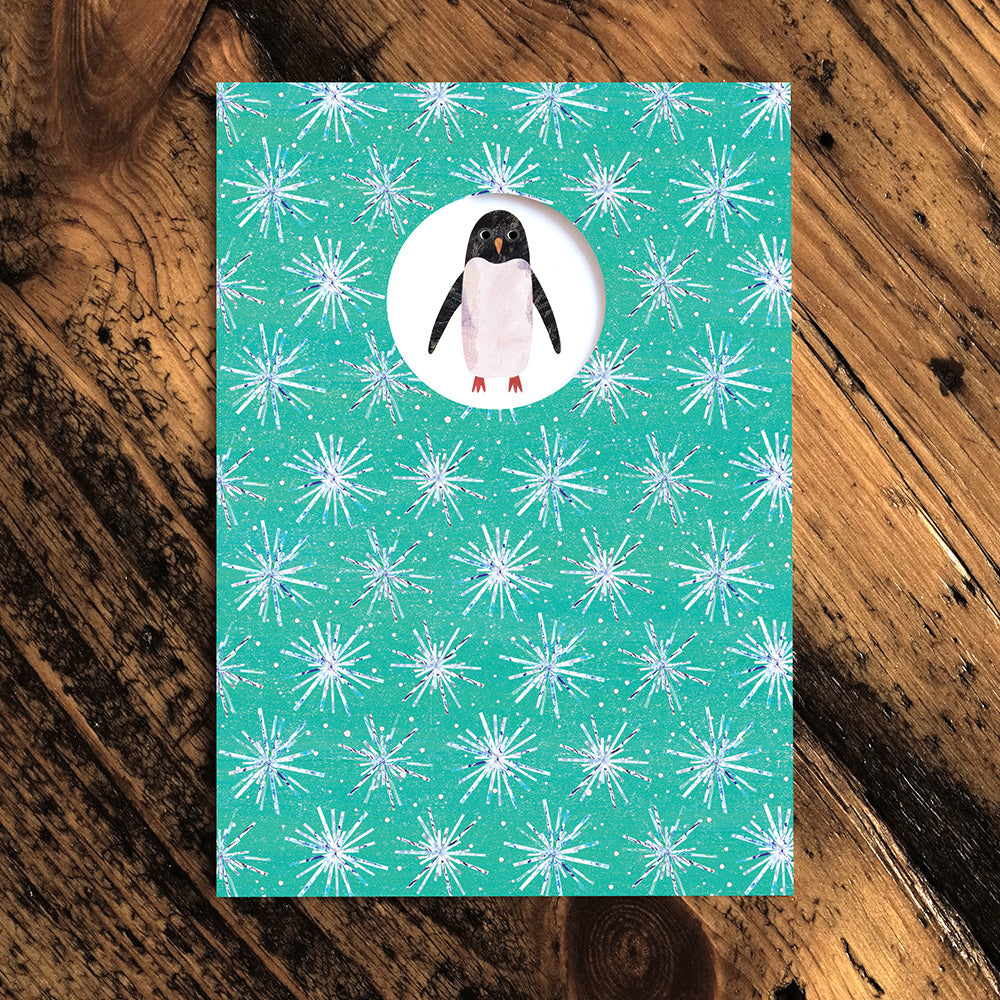 Christmas card with a pale green background and white snowflake pattern. White circle cut out with a penguin illustration. 