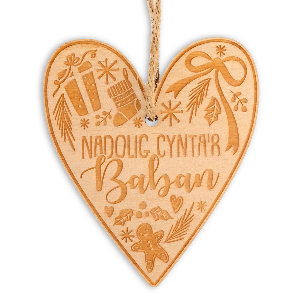 Heart shaped wooden laser cut hanging Christmas decoration. The text in the centre says 'Nadolig Cynta'r Baban' it is surrounded by festive foliage, bows, gingerbread, stockings, snowflakes, hearts and presents. 
