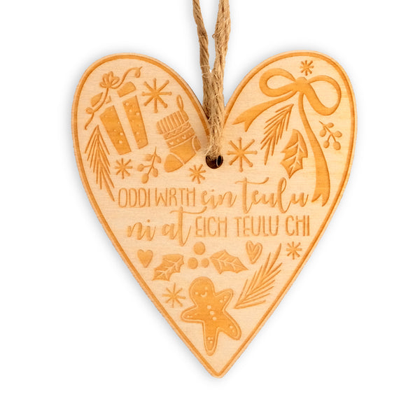 Heart shaped wooden laser cut hanging decoration with jute twine. The design has 'Oddi wrth ein Teulu ni at eich Teulu chi' in the centre, with the surrounding edges full of festive foliage, stars, hearts and ribbons, gingerbreadmen, stockings and presents.  