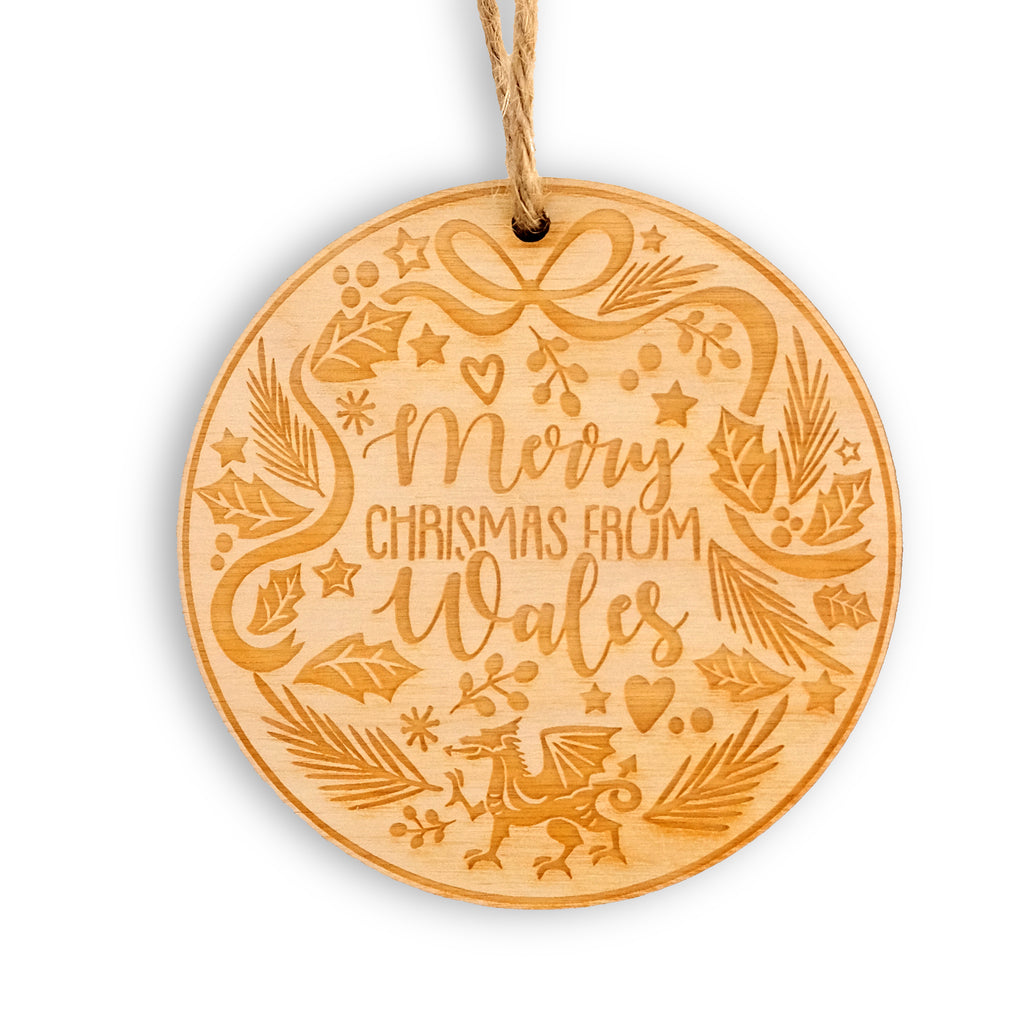 Round wooden laser cut hanging decoration with jute twine. The design has 'Merry Christmas from Wales' in the centre, with the Welsh dragon at the bottom with a wreath style design around the edge of festive foliage, stars, hearts and ribbons. 