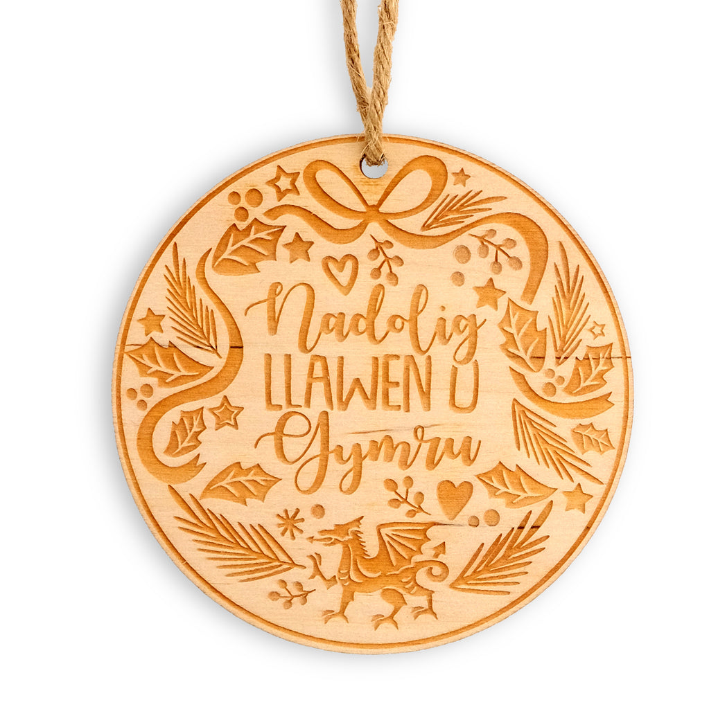 Round wooden laser cut hanging decoration with jute twine. The design has 'Nadolig Llawen o Gymru' in the centre, with the Welsh dragon at the bottom with a wreath style design around the edge of festive foliage, stars, hearts and ribbons. 