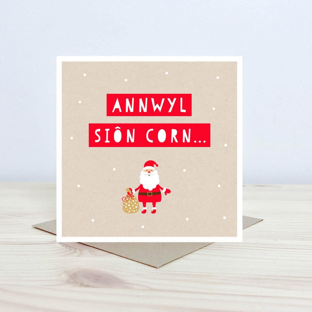 Welsh language greeting card with a cartoon style illustration of Father Christmas carrying a sack on a kraft background with snowballs falling. Text is across the top in white on a red background and reads "Annwyl Sion Corn..."