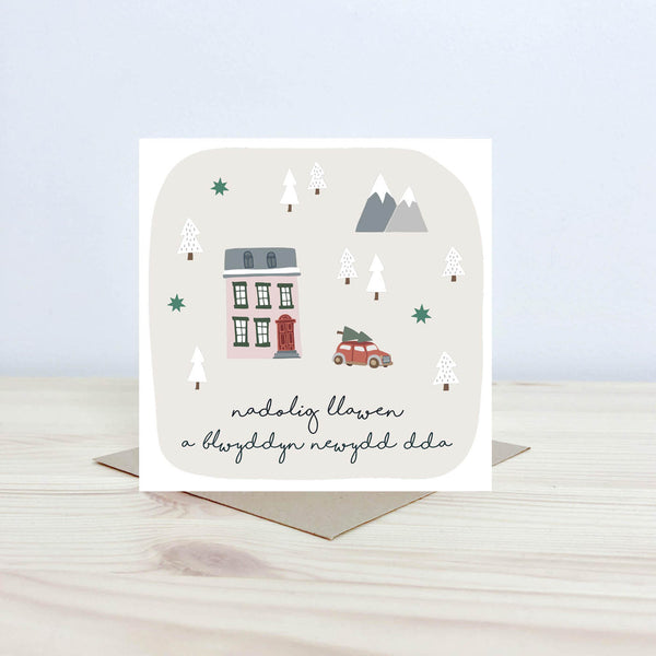 Welsh language Christmas card featuring the text 'Nadolig Llawen a Blwyddyn Newydd Dda' the illustration shows a house with a car parked by with a christmas tree attached to its roof. There are snowcapped tree and mountains in the background