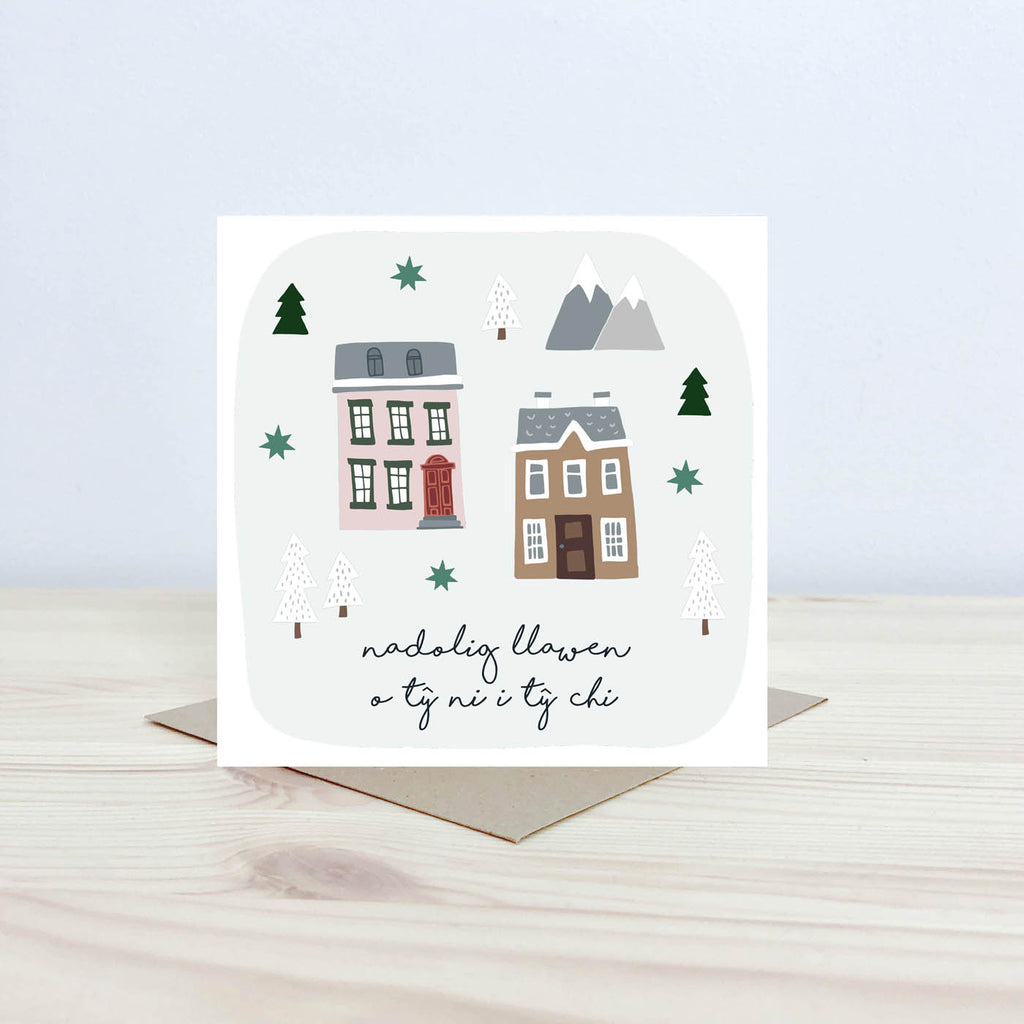 Welsh language greeting card featuring the text 'Nadolig Llawen o Ty ni i ty chi'. Two illustrations of houses next to each other with snowcapped trees and mountains in the background.