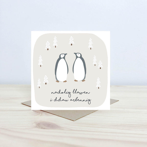 Welsh language greeting card with the text 'Nadolig Llawen i ddau arbennig' featuring two penguins looking at each other surrounded by snowcapped christmas trees. 
