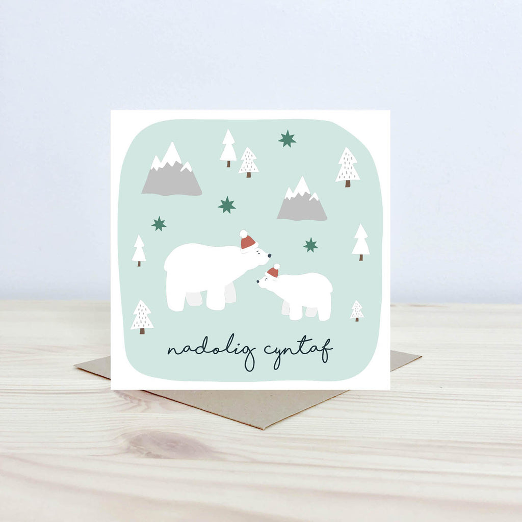 Welsh language Christmas card featuring a large parent polar bear and a child polar bear both wearing christmas hats. On a pale blue background surrounded by snow covered trees and mountains.