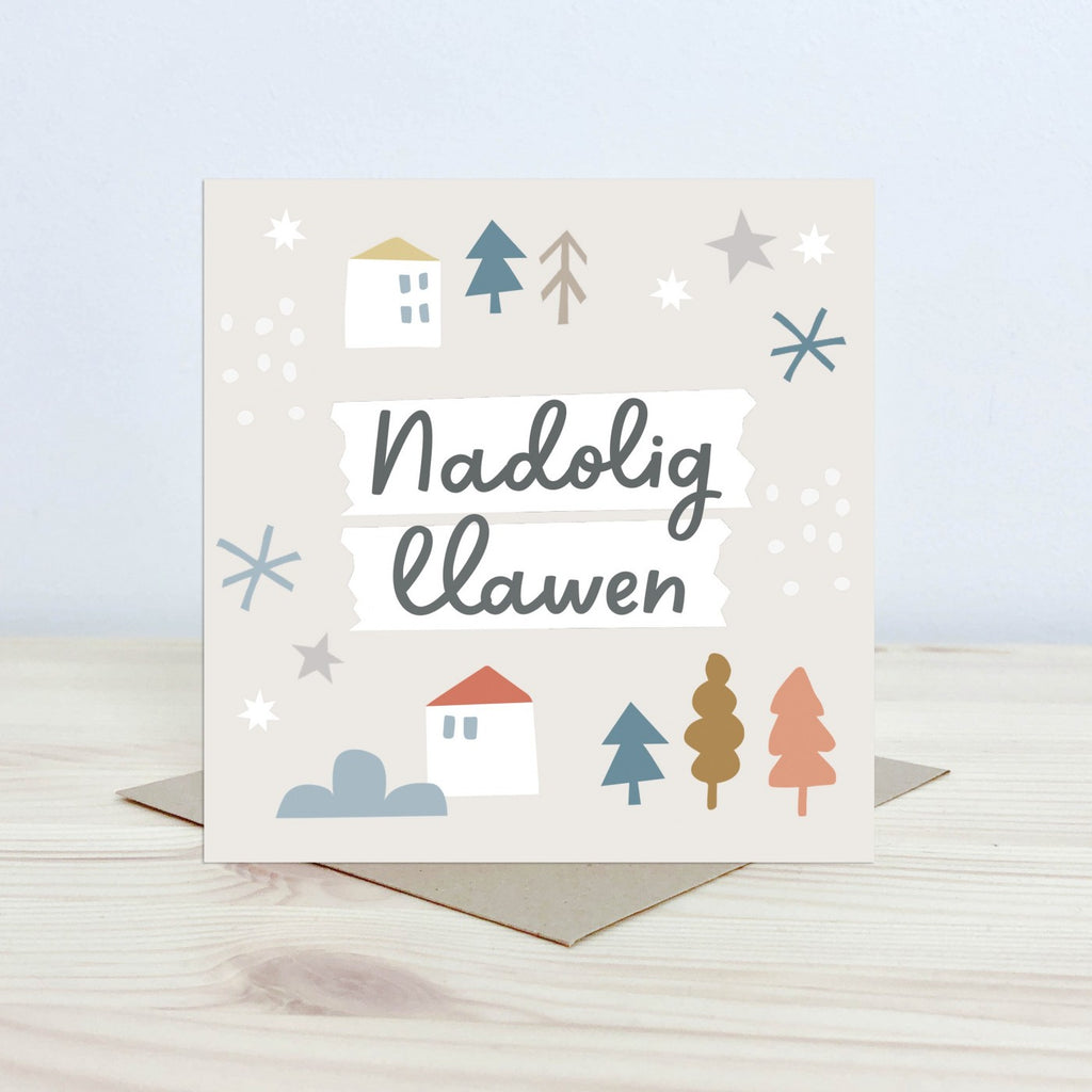 Christmas Card with the text 'Nadolig Llawen' featured on the front in black cursive font on a white background. The background is pale cream with graphic block houses, stars, snowflake and tree shapes. 