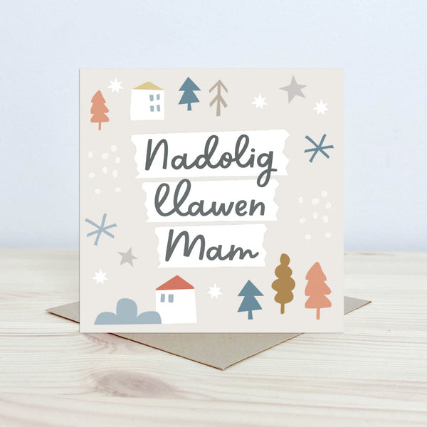 Christmas Card with the text 'Nadolig Llawen Mam' featured on the front in black cursive font on a white background. The background is pale cream with graphic block houses, stars, snowflake and tree shapes. 