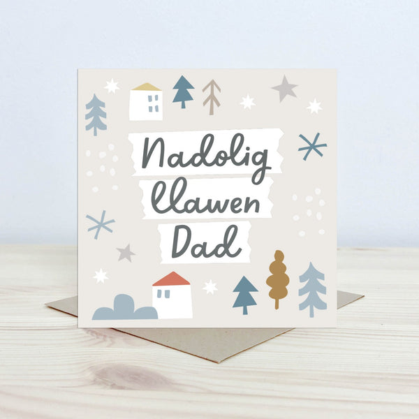 Christmas Card with the text 'Nadolig Llawen Dad' featured on the front in black cursive font on a white background. The background is pale cream with graphic block houses, stars, snowflake and tree shapes. 