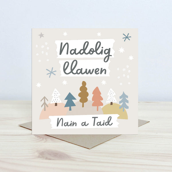 Christmas Card with the text 'Nadolig Llawen Nain a Taid' featured on the front in black cursive font on a white background. The background is pale cream with graphic stars, snowflake and tree shapes. 