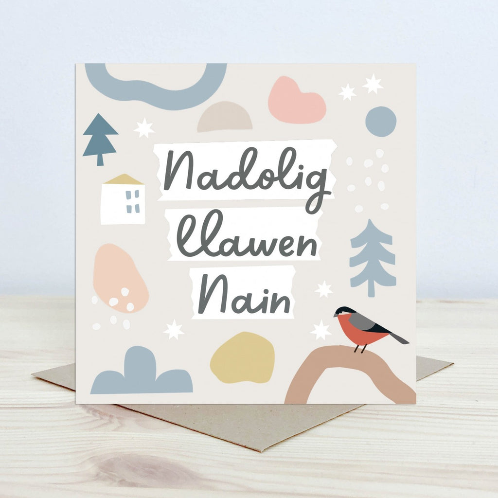 Christmas Card with the text 'Nadolig Llawen Nain' featured on the front in black cursive font on a white background. The background is pale cream with a bird on a branch in the foreground and graphic stars, snowflake and tree shapes. 