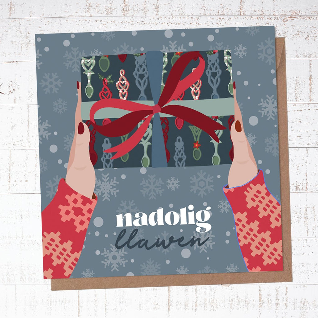 Greeting card with an illustration of hands in a christmas jumper hanging over a present wrapped in festive paper decorated in lovespoons with a red bow. 