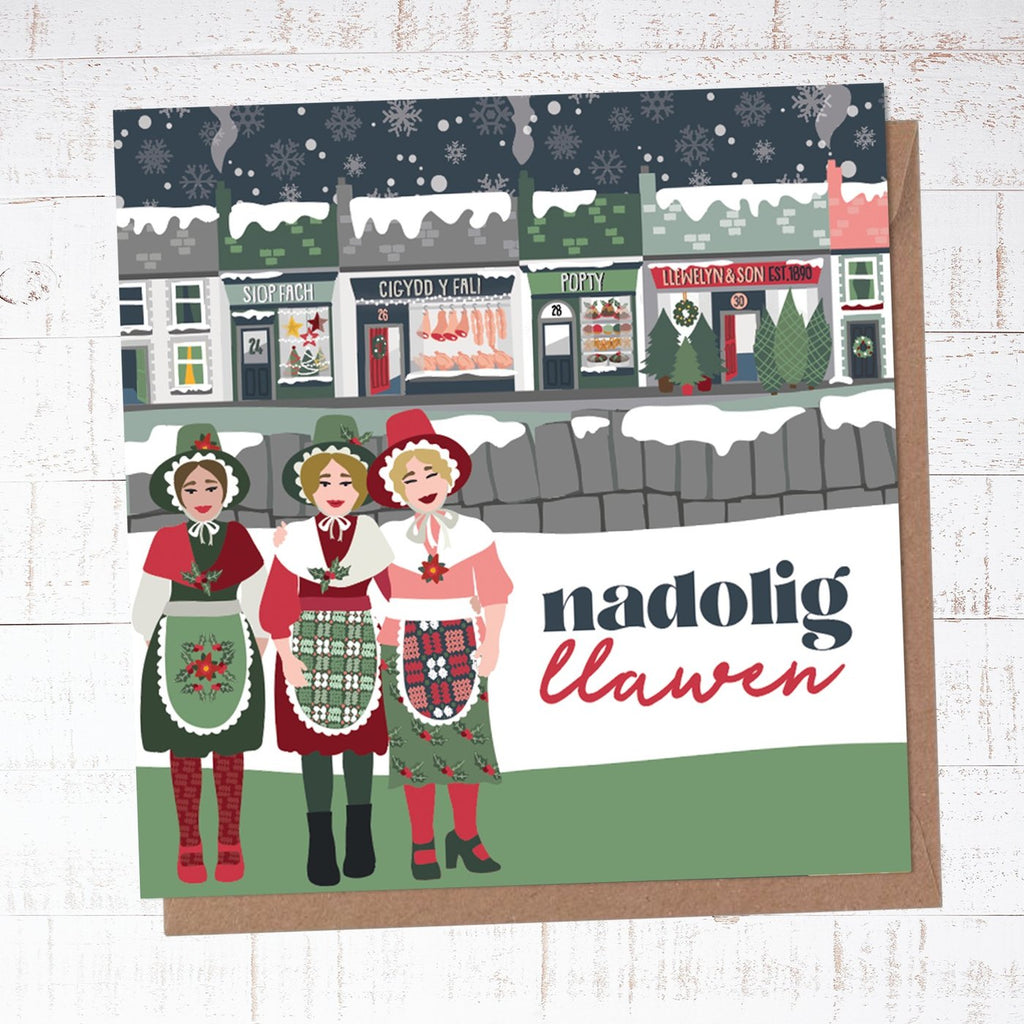 Christmas card with 'Nadolig Llawen'. Three welsh ladies stood in the foreground wearing festive traditional Welsh costume. Behind them is a snow covered stone wall and a highstreet scene of snow covered shops. 
