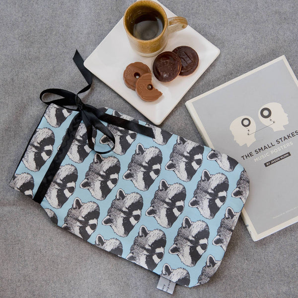 Lifestyle image.  Blue fabric print fabric with raccoon motif hot water bottle.  Black ribbon.