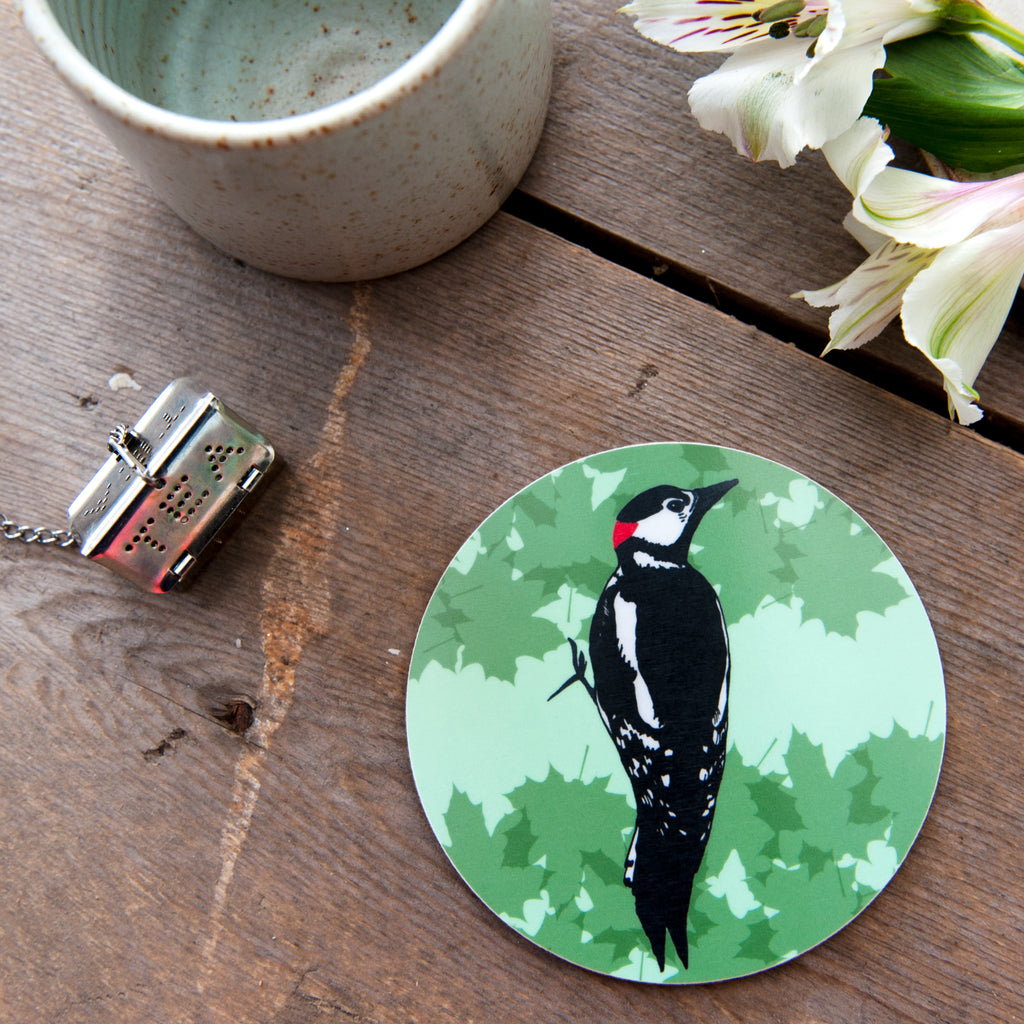 Lifestyle flatlay photograph of a wooden table with an empty ceramic mug and tea strainer and some white flowers. The main focus in a round coaster with an illustrated woodpecker and green leaf background. 