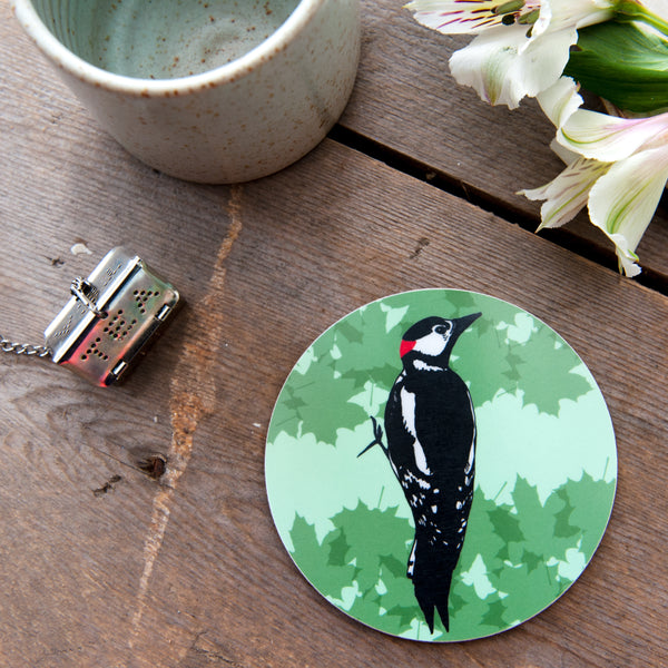 Lifestyle flatlay photograph of a wooden table with an empty ceramic mug and tea strainer and some white flowers. The main focus in a round coaster with an illustrated woodpecker and green leaf background. 