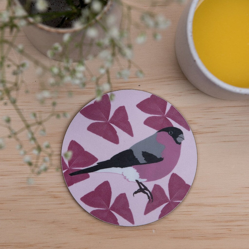 Lifestyle photo showing a round coaster on a table next to flowers and a mug. The coaster is round with a pink floral pattern and an illustration of a bullfinch.